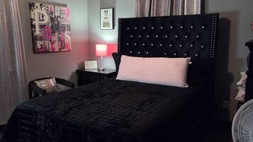 Une chambre à coucher avec une tête de lit capitonnée, un lit avec un oreiller rose, une table de chevet avec une lampe, une horloge murale et une affiche.