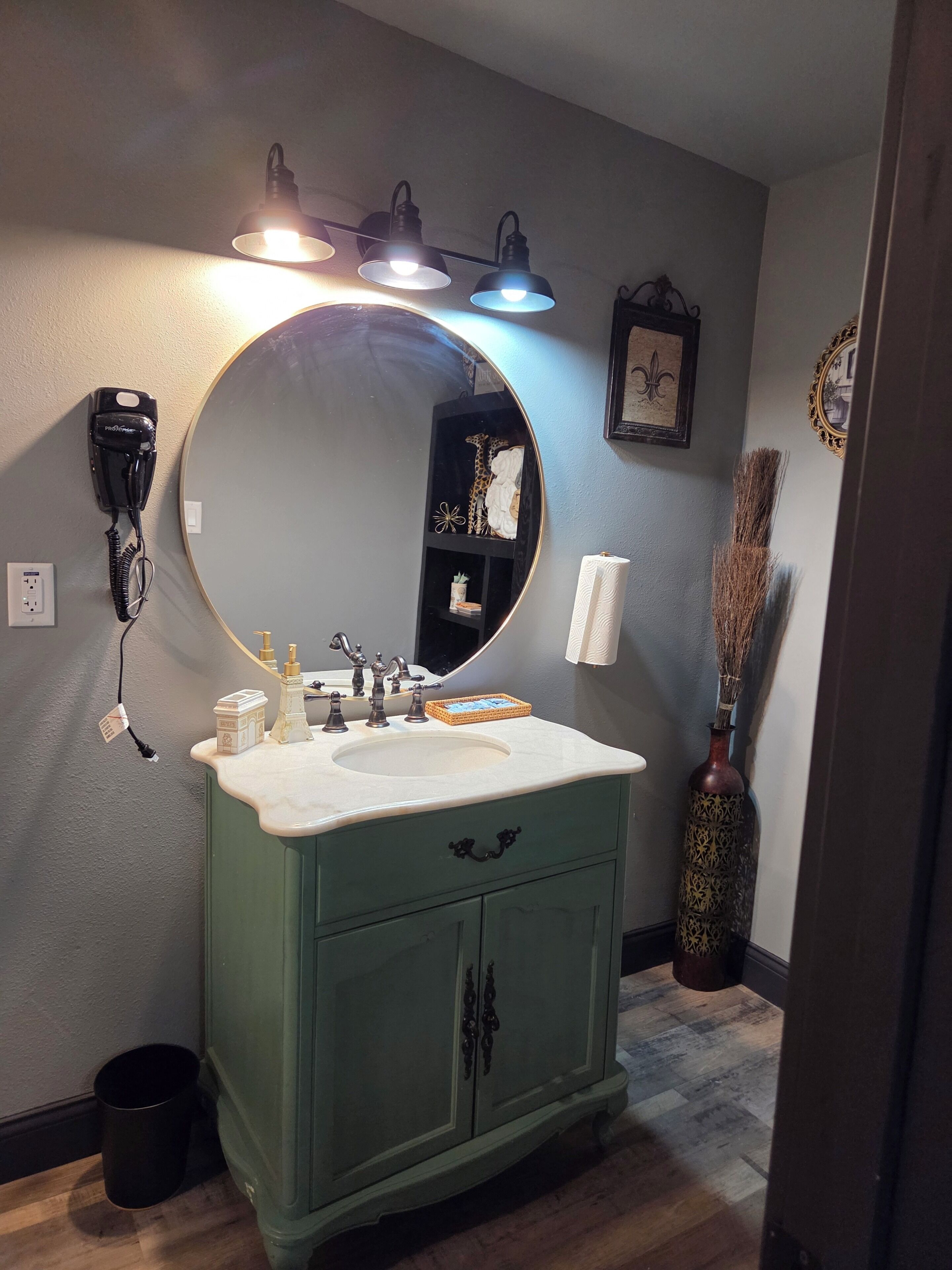Une salle de bain équipée d’un meuble-lavabo, d’un miroir rond, d’un sèche-cheveux, d’un lavabo et d’un vase.