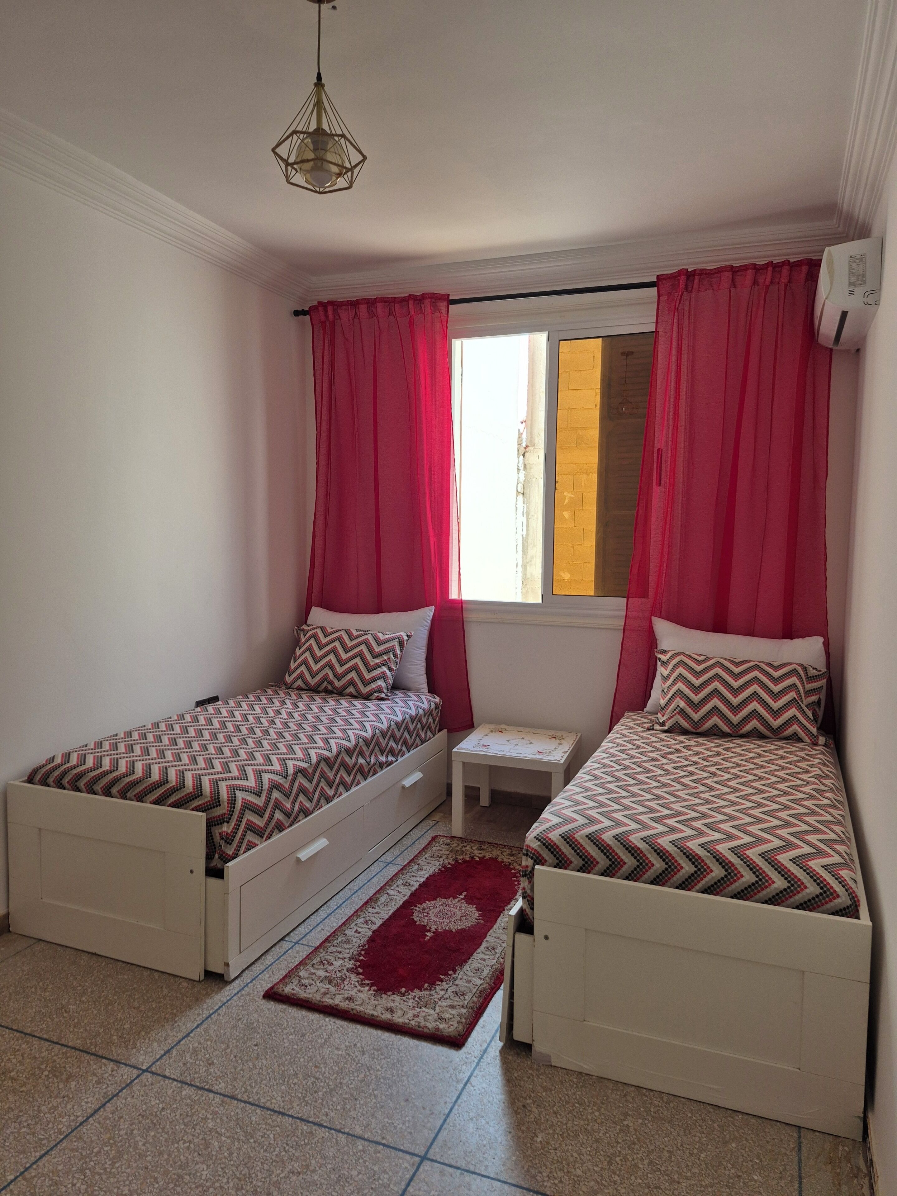 A bedroom with two single beds, a small table, a window with curtains, and a hanging light fixture.