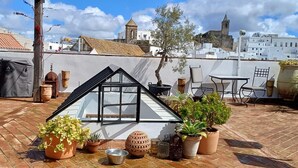 Una terraza en la azotea con una estructura de techo de vidrio, plantas en macetas, mobiliario exterior y vistas a edificios históricos.