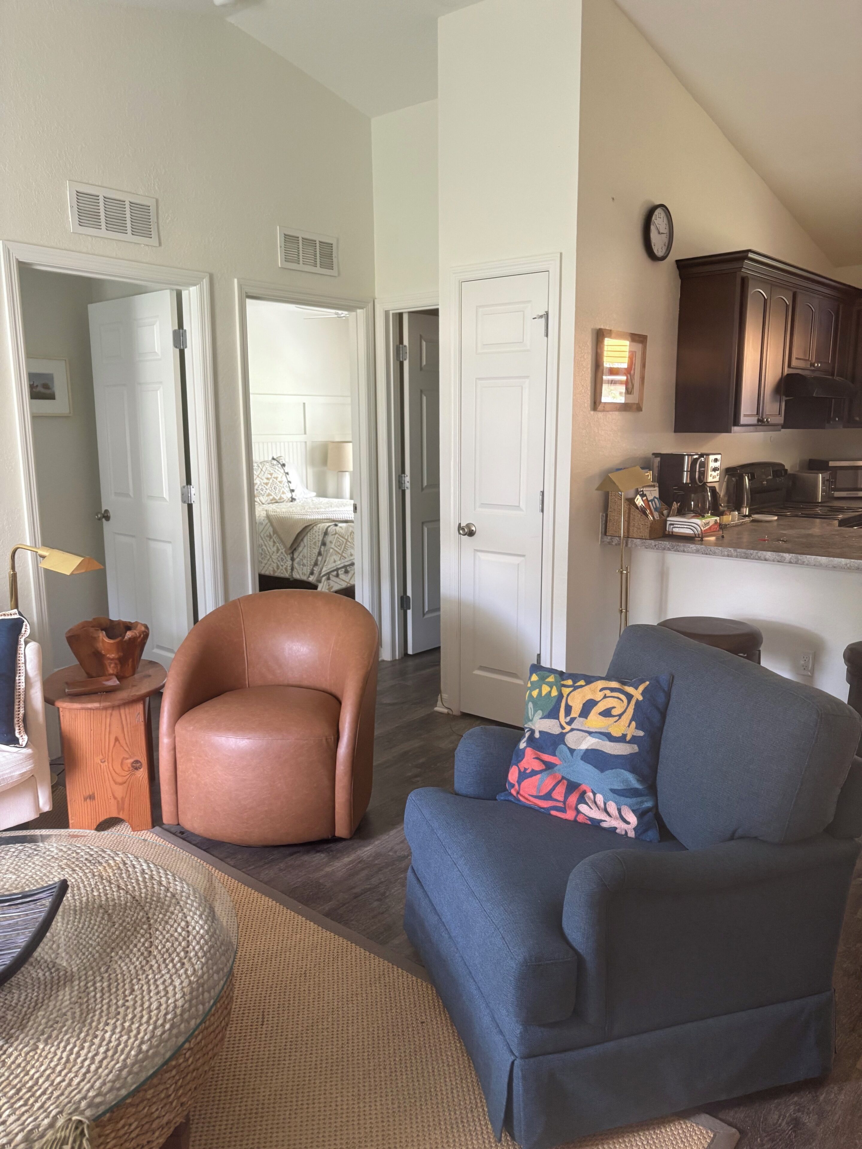 A living room with a brown leather chair, a blue sofa with a colorful pillow, a woven basket, and a kitchen area with cabinets and appliances.