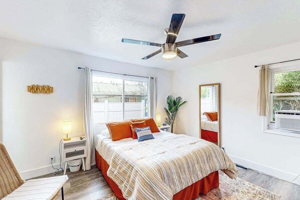 A bedroom with a bed, a ceiling fan, a window, a nightstand, and a plant.