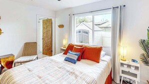Une chambre à coucher avec un lit, une chaise, une table de chevet, une lampe et une fenêtre avec des rideaux.