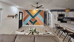 A modern living room with a sofa, a dining area with a table and chairs, a kitchen with white cabinets, and a decorative wall with geometric patterns. - New Dog-Friendly Pahoa Home with Yard, 31-Night Minimum (Pahoa)