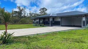 A modern two-story house with a garage, surrounded by a well-maintained lawn and a few trees. - New Dog-Friendly Pahoa Home with Yard, 31-Night Minimum (Pahoa)