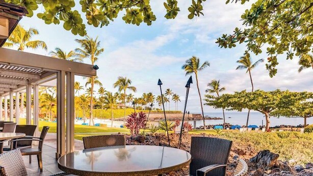 An outdoor seating area with wicker furniture, a covered section, and a view of palm trees and a body of water. - NEW Mauna Lani Space, Pool Time, and Kohala Coast Sun (Waimea)