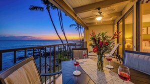 A balcony with a table set for two, two wine glasses, a vase with flowers, and a view of the ocean at sunset. - *NEW* Oceanfront Kona Retreat Near Lyman's Surf Spot (Kailua-Kona)