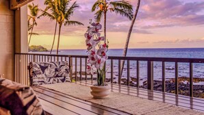 A wooden deck with a potted orchid, a patterned sofa, and a view of the ocean with palm trees. - NEW Fall Deal Wide Pacific Views from a Two-Bedroom Kona Condo (Kailua-Kona)