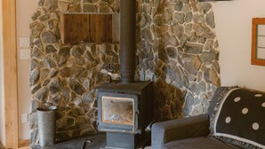 A stone wall with a wood-burning stove, a wooden ceiling, and a gray sofa with a polka dot throw. - Rustic Lakefront Lodge on Little Atlin Lake - Pet friendly! (Tagish)
