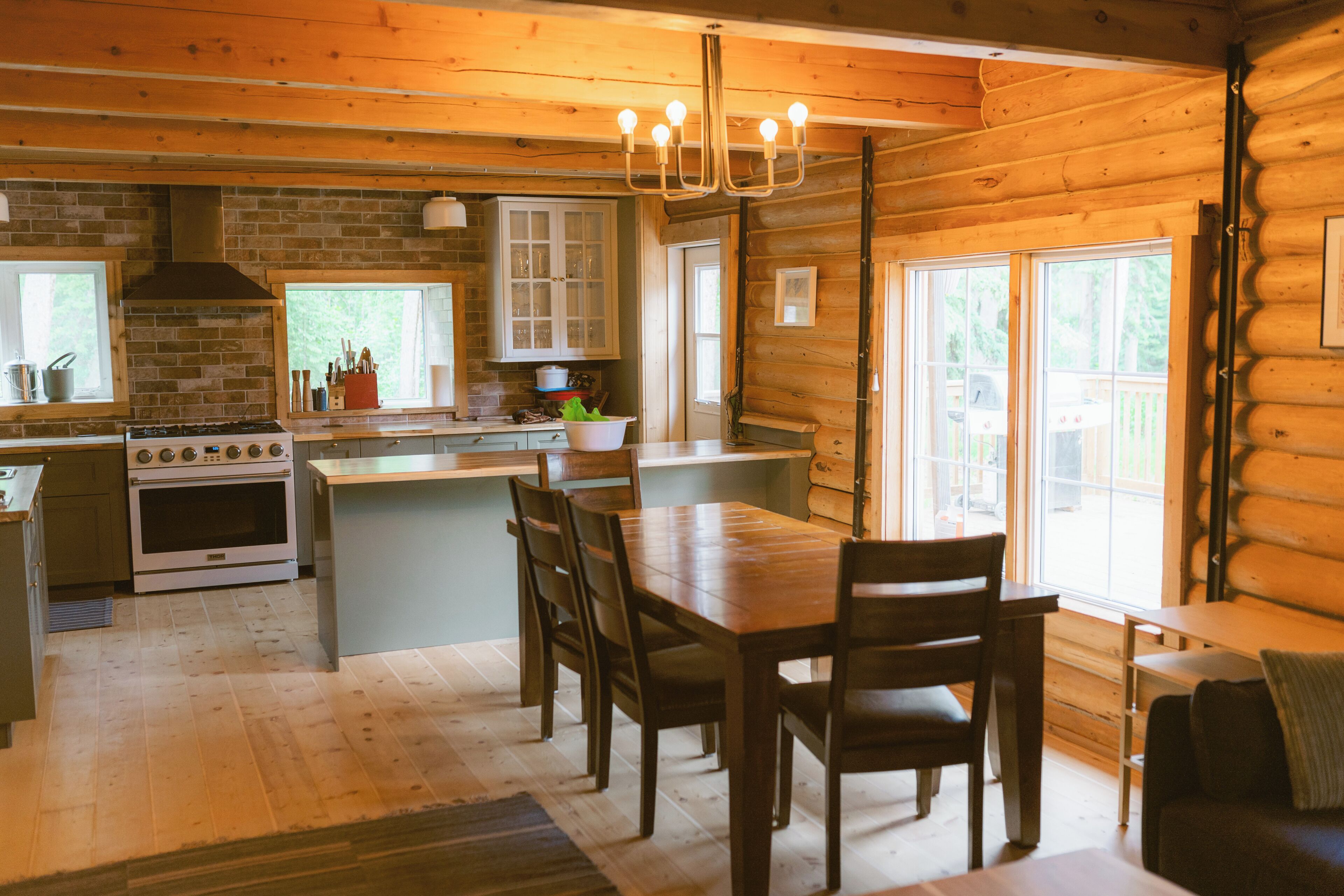 A wooden cabin interior with a kitchen, dining area, and large windows.