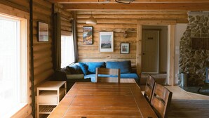 A wooden cabin interior with a dining table, blue sofa, and a stone fireplace. - Rustic Lakefront Lodge on Little Atlin Lake - Pet friendly! (Tagish)