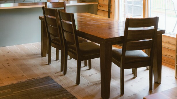 A wooden interior with a dining table, chairs, a chandelier, and a kitchen area with cabinets and a window. - Rustic Lakefront Lodge on Little Atlin Lake - Pet friendly! (Tagish)