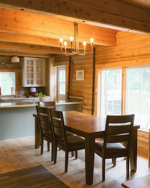 A wooden interior with a dining table, chairs, a chandelier, and a kitchen area with cabinets and a window.