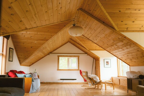 An attic room with wooden beams, a sloped ceiling, a window, a bed, a chair, and a radiator.