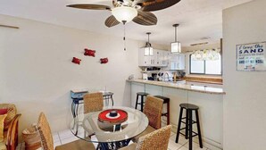 A dining area with a glass table, wicker chairs, a ceiling fan, and a kitchen in the background. - *New* Fall Special Kona Family Condo Near Beach, Pool and Hot Tub (Kailua-Kona)