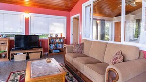 Un salon avec un plafond en bois, un canapé beige, un téléviseur, une bibliothèque et une fenêtre avec des stores.