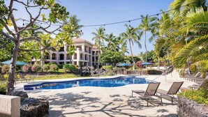 A pool area with lounge chairs, palm trees, and a building with arched windows. - *New* Fall Sale Renovated Waikoloa Condo Near A-Bay (Waikoloa)