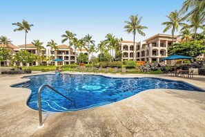 A swimming pool with a curved edge, surrounded by palm trees and a building with multiple balconies. - *New* Fall Sale Renovated Waikoloa Condo Near A-Bay (Waikoloa)