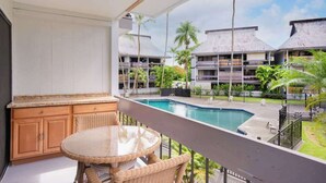A balcony with a view of a swimming pool, wooden furniture, and a building with multiple balconies. - *New* Fall Sale Hilo Water-View Condo with Lanai, Pool, and Koi Stream (Hilo)