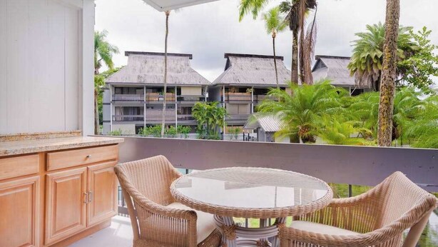 A balcony with wicker furniture, a glass table, and a view of a resort with multiple buildings and palm trees. - *New* Fall Sale Hilo Water-View Condo with Lanai, Pool, and Koi Stream (Hilo)