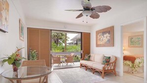 A room with a glass table, wicker furniture, a ceiling fan, and a view of greenery outside. - *New* Fall Sale Hilo Water-View Condo with Lanai, Pool, and Koi Stream (Hilo)