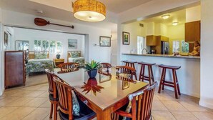 Un espace repas avec une table et des chaises en bois, un luminaire suspendu et une cuisine visible à travers une porte ouverte.