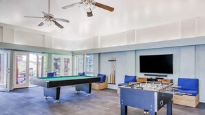 Une table de billard, une table de baby-foot et un téléviseur dans une pièce dotée de grandes fenêtres et de ventilateurs de plafond.
