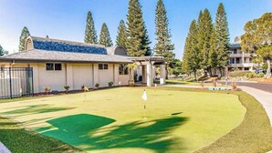 Un parcours de golf avec un green d’entraînement, un club-house et de grands arbres en arrière-plan.