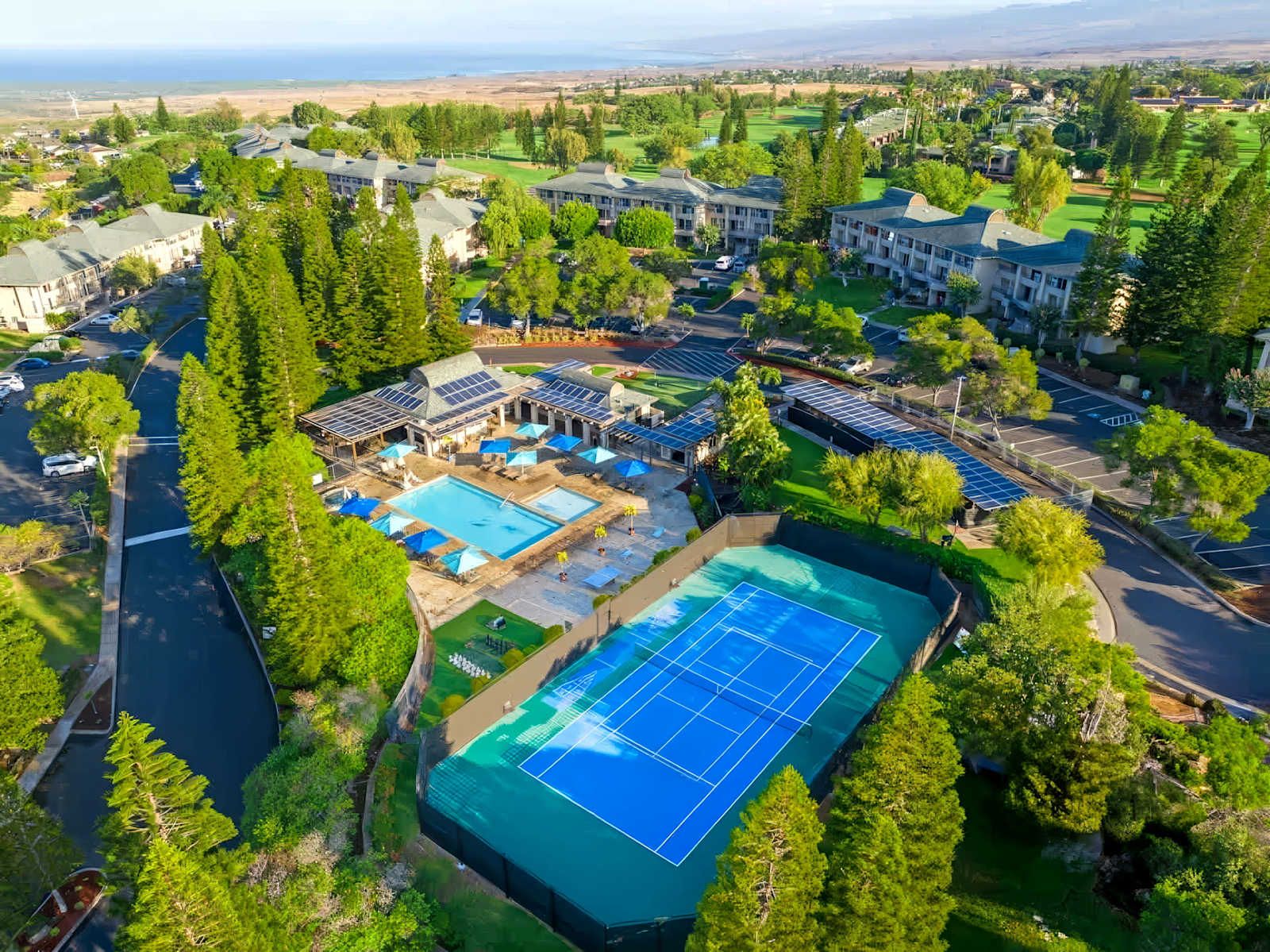 An aerial view of a resort with multiple buildings, a swimming pool, tennis courts, and solar panels.