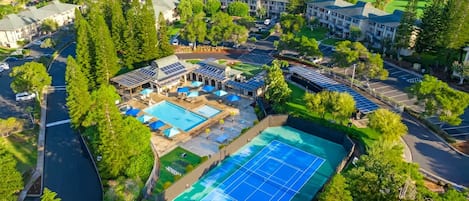 An aerial view of a resort with multiple buildings, a swimming pool, tennis courts, and solar panels.