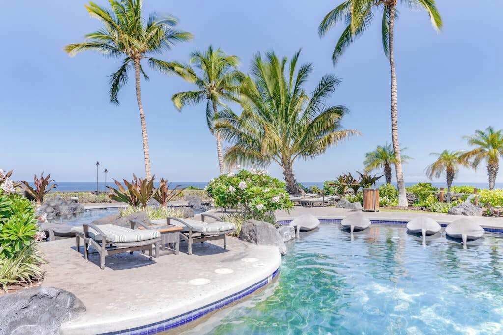 A pool area with lounge chairs, palm trees, and a view of the ocean.