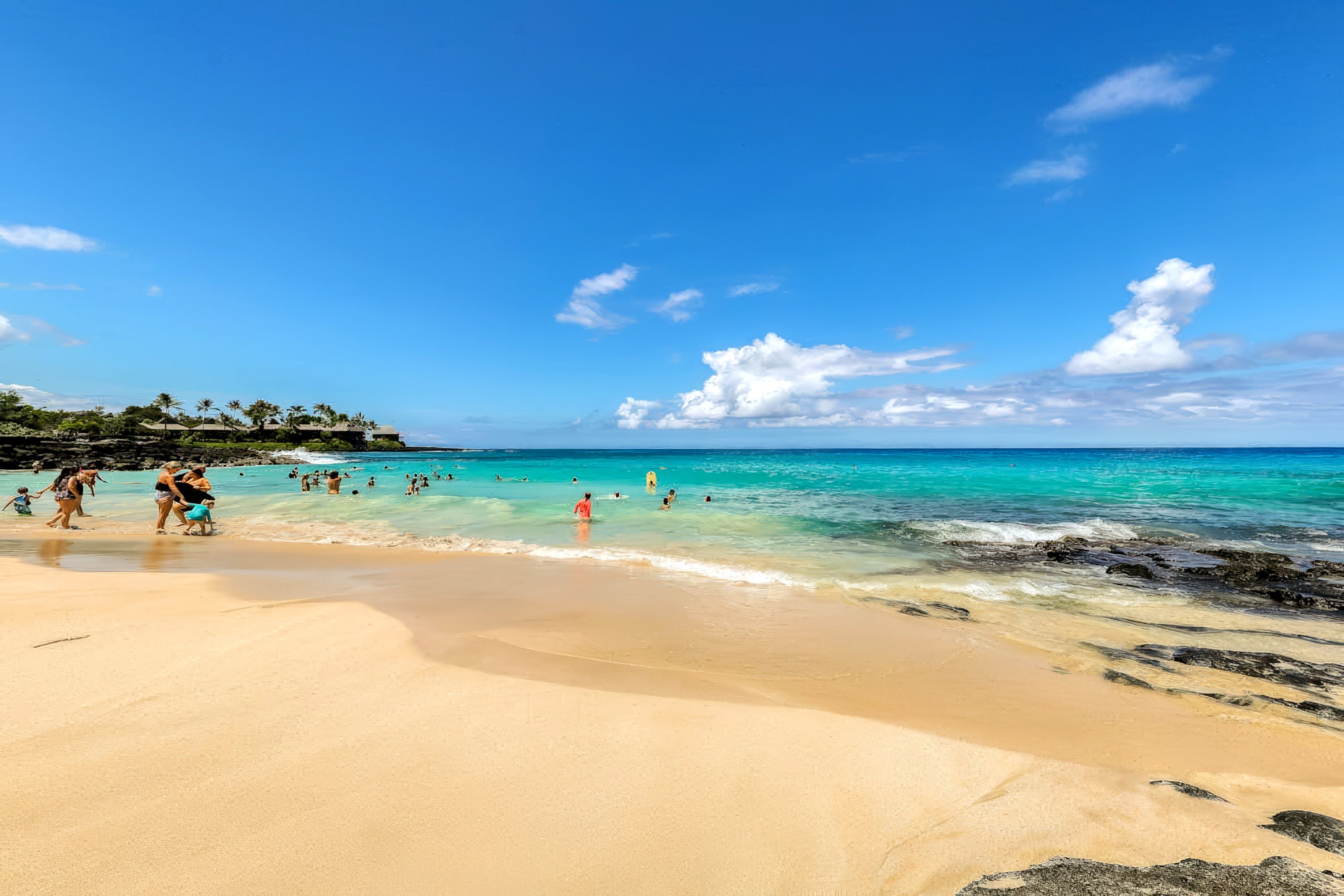 Une plage avec une eau turquoise et limpide, un rivage sablonneux et quelques personnes qui se baignent.