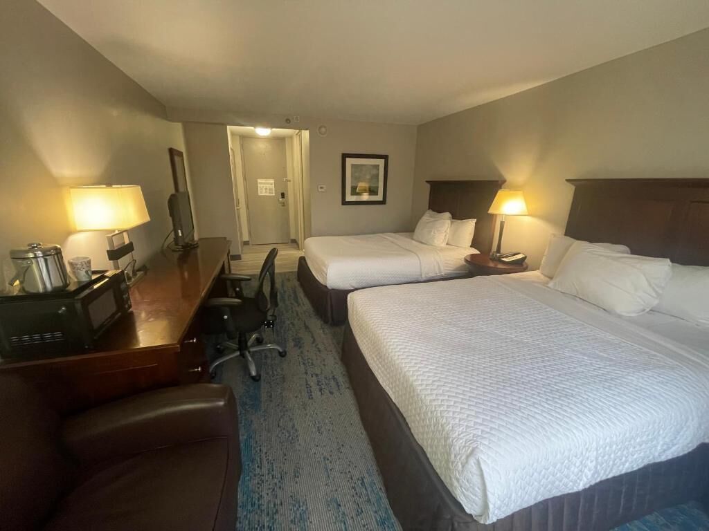 Habitación de hotel con dos camas, un escritorio con lámpara, una silla y una pequeña mesa con lámpara.