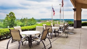 An outdoor patio with round tables and chairs, American flags, and a view of greenery and buildings in the distance.