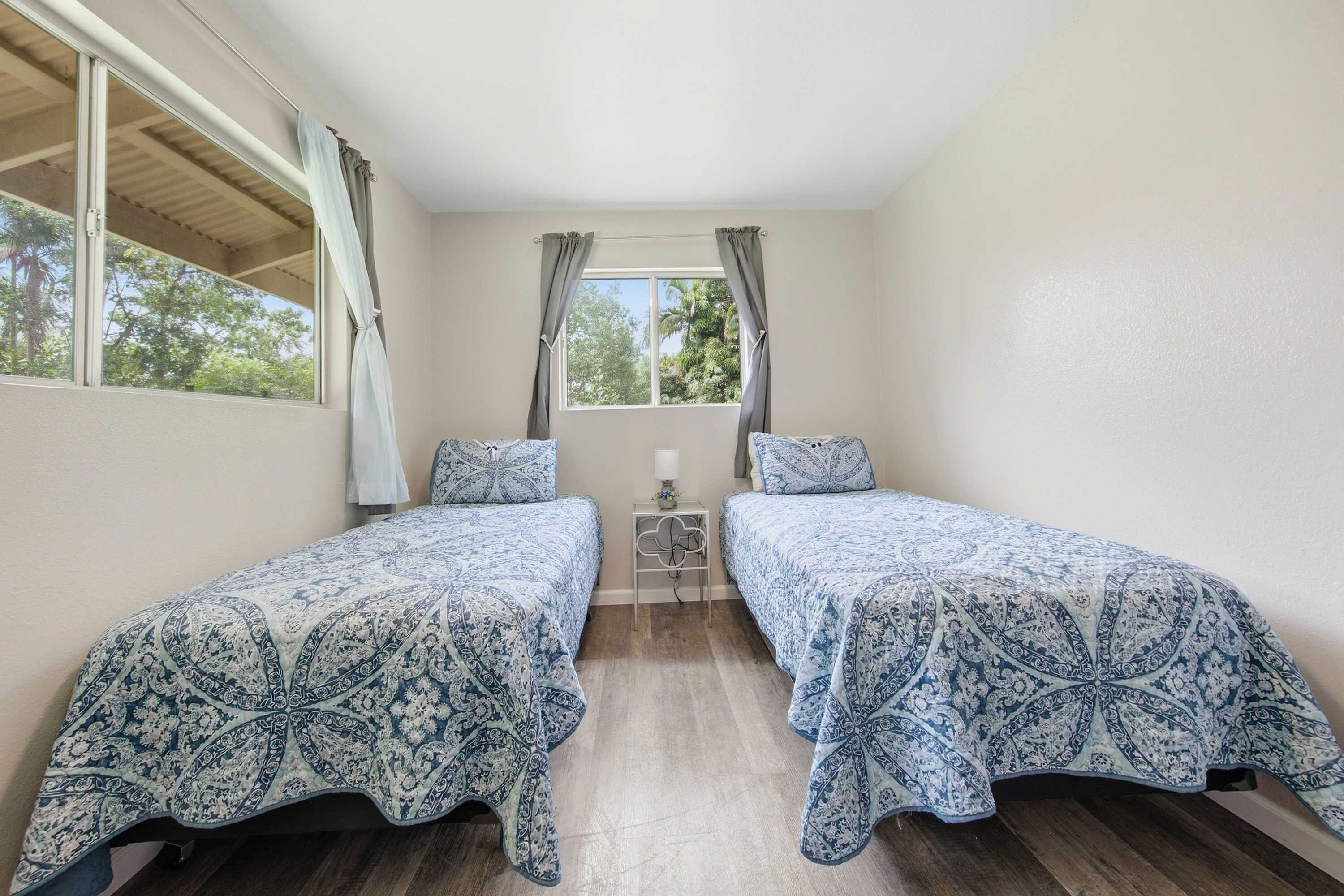 A bedroom with two single beds, a small bedside table, a window with curtains, and a view of trees.