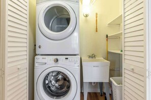 A compact laundry area with a stacked washer and dryer, a sink, and a trash bin. - Kona Town Mansions Beachside Condo w/ Pool, AC, BBQ, Kitchen- Walk to Activities (Kailua-Kona)