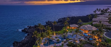 Un resort con piscina, zonas de descanso y vista al océano al atardecer.