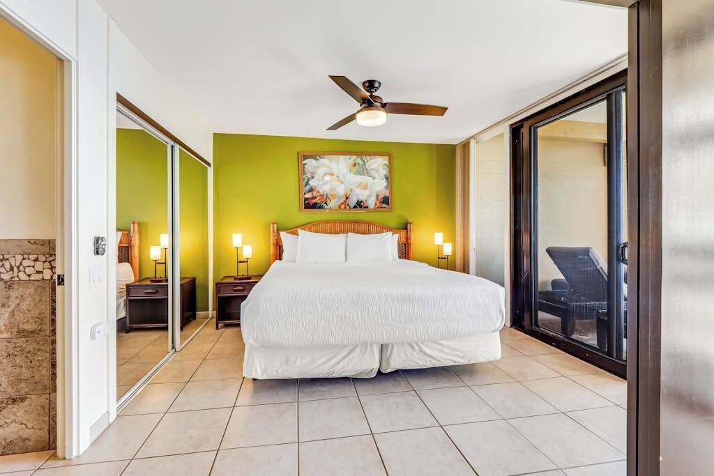 A bedroom with a large bed, a ceiling fan, a painting on the wall, and a sliding glass door leading to a balcony.