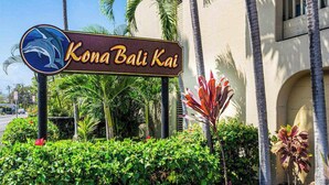 Schild für Kona Bali Kai mit einem Delphin-Logo, umgeben von tropischen Pflanzen und Palmen.
