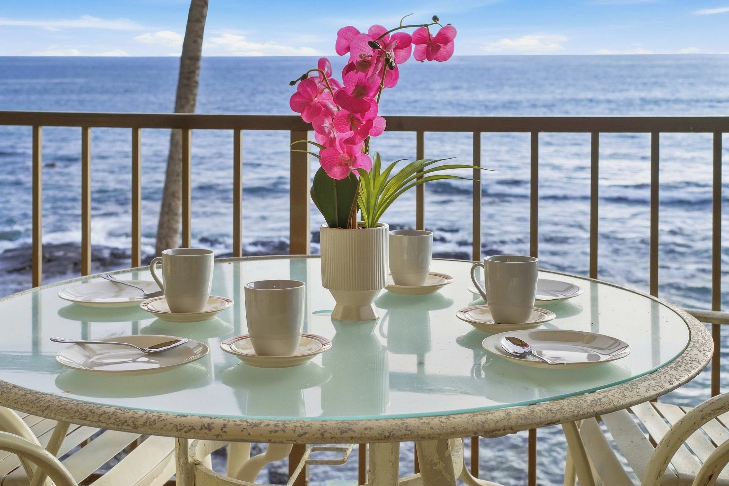Une table en verre, ornée d’assiettes, de tasses et d’un vase rempli de fleurs roses, le tout placé devant une vue sur l’océan.