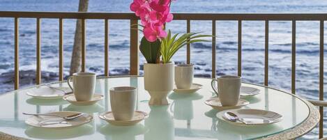 A glass table with plates, cups, and a vase of pink flowers, set against a view of the ocean.