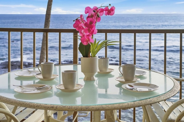 Una mesa de vidrio con platos, tazas y un florero de flores rosas, con vista al océano.
