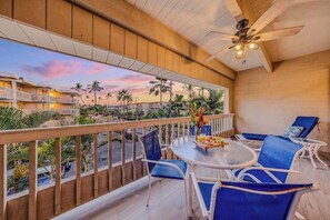 A balcony with a view of palm trees and a sunset, furnished with blue chairs and a round table. - Kona Beach Condo with Shared Pool, Hot Tub & Private Lanai (Kailua-Kona)