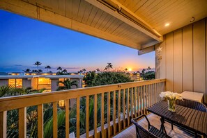 Un balcon offrant une vue sur un coucher de soleil sur une zone côtière, avec une balustrade en bois, une table en osier agrémentée d’un vase de fleurs et un petit coin salon.