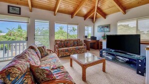 Un salón con techo de madera, un sofá, una mesa de centro, un televisor y ventanas grandes con vistas a palmeras.