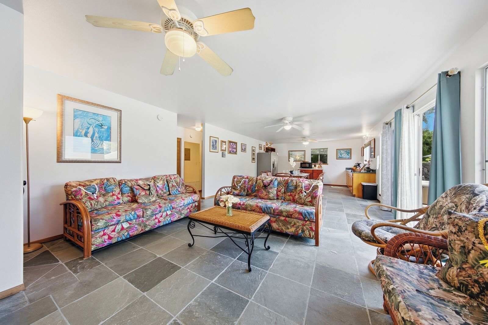 A spacious living area with a ceiling fan, two sofas with floral upholstery, a coffee table, and a dining area with a kitchen in the background.