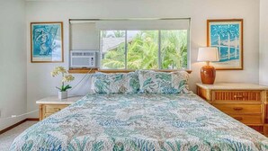 Un dormitorio con cama, ventana, aire acondicionado, mesita de noche, lámpara y una obra de arte.