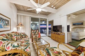 Un salon doté d’un ventilateur de plafond, d’un canapé à motifs floraux, d’un téléviseur posé sur un meuble en bois et d’un lit recouvert d’une couverture blanche.
