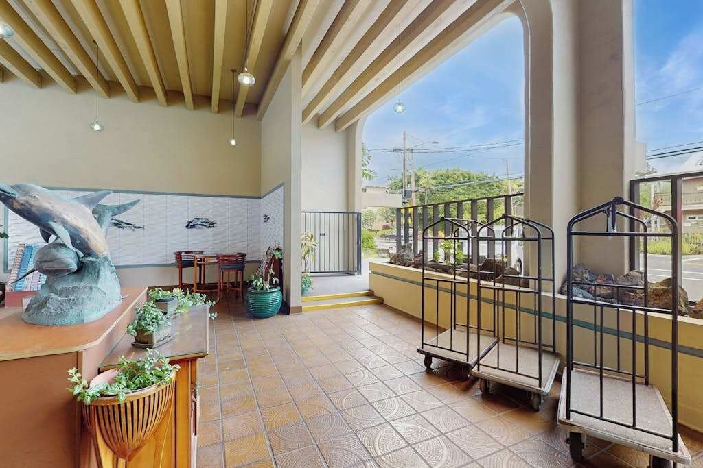 A spacious lobby with a tiled floor, a reception desk with a sculpture, potted plants, and a view through a large window.