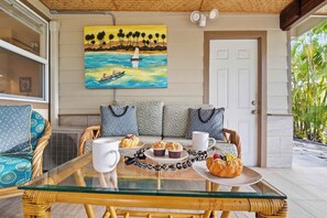 A dining area with a glass table, wicker chairs, a sofa with patterned cushions, a painting of a tropical scene, and a door leading to a covered outdoor space. - Kailua View Estates - Private Home Retreat w/ Lanai, Hot Tub, Laundry, BBQ, A/C (Kailua-Kona)